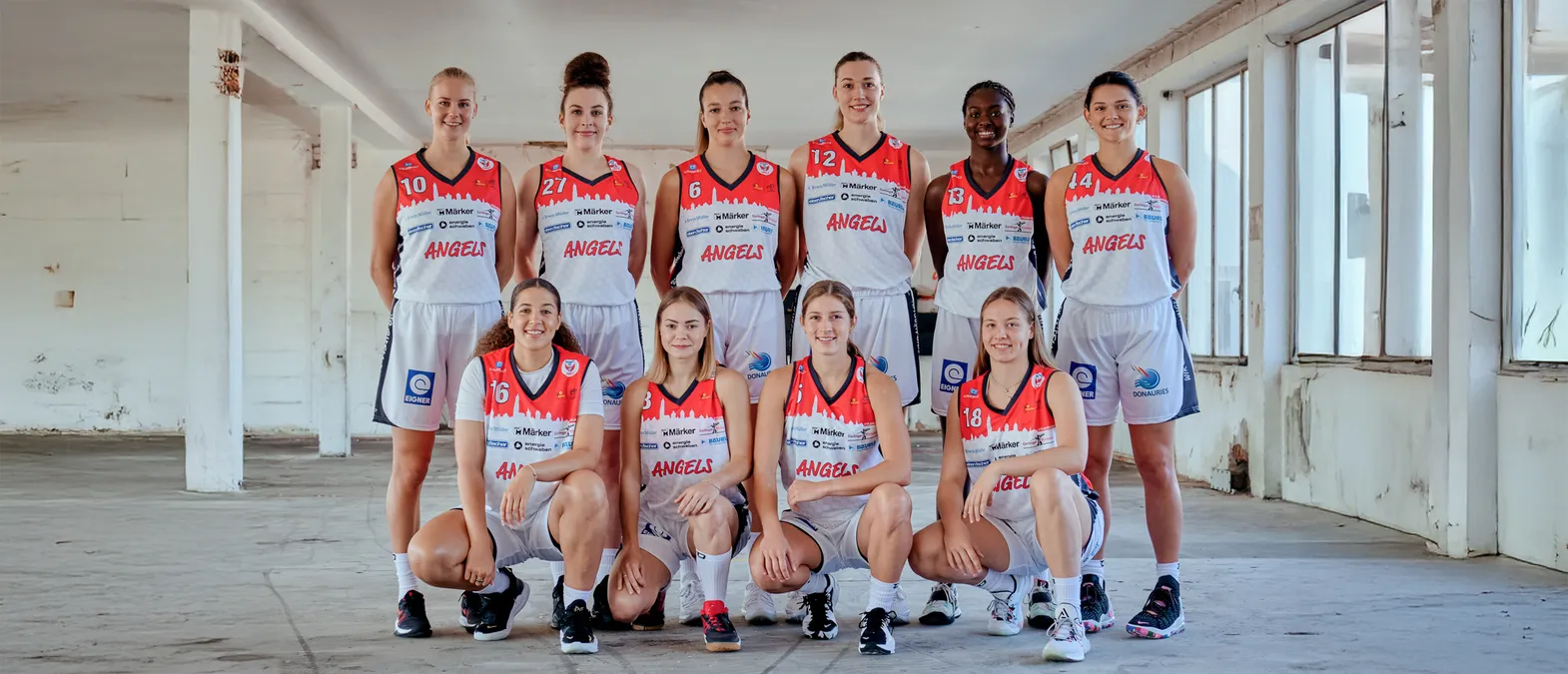 Un grupo de ocho mujeres con uniformes de baloncesto naranja y blanco con la palabra 'ANGELS' posan en un almacén.