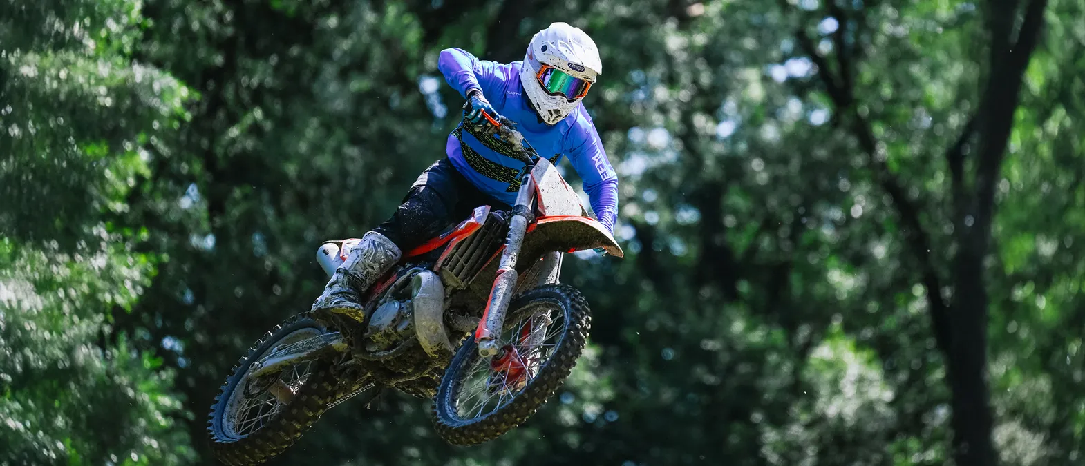 Un motociclista con un traje de carreras azul de manga larga y casco blanco salta en una motocicleta a través de un bosque.