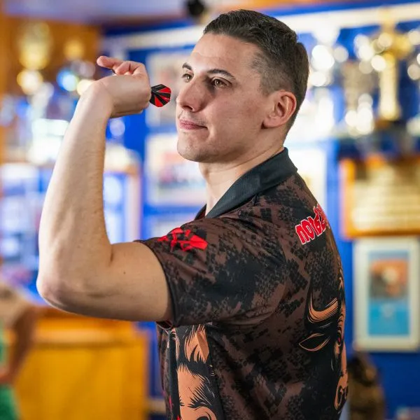 Un hombre con una camiseta deportiva estampada con detalles rojos sostiene un dardo y apunta, con trofeos visibles en el fondo.