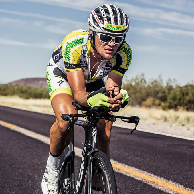 Un ciclista con un maillot amarillo y negro y guantes conduce en una carretera, usando un casco de ciclismo y gafas de sol.