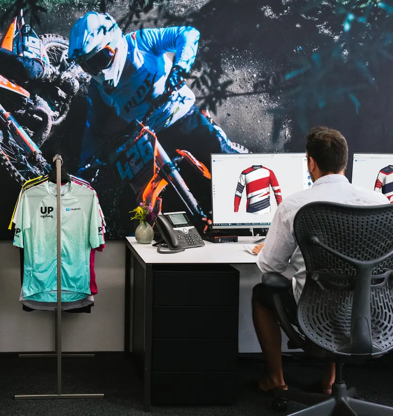 Un hombre está sentado en un escritorio diseñando una camiseta deportiva personalizada en una computadora, mientras que en el fondo se ve una gran imagen de un motociclista y un perchero con varias prendas deportivas.