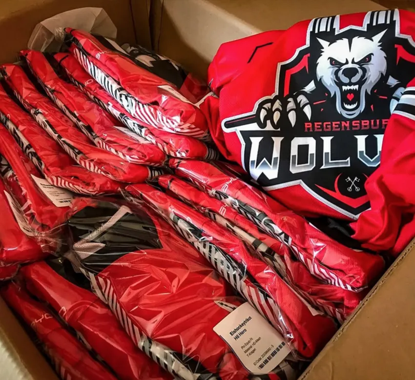 Una caja de cartón llena de varias camisetas deportivas rojas personalizadas envueltas en plástico, una de ellas con un logo de lobo y la inscripción 'REGENSBURG WOLVES'.