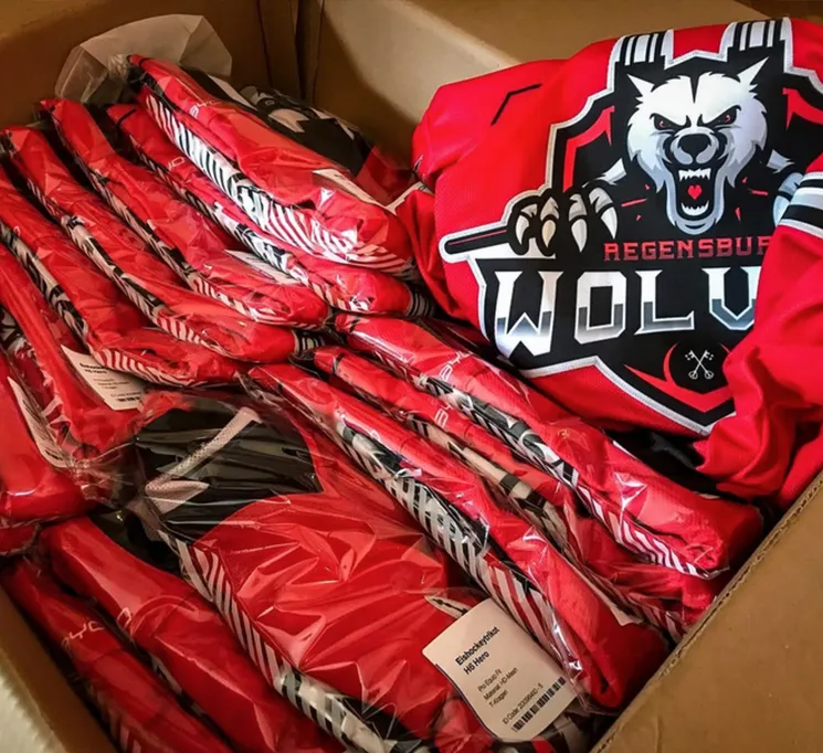 Una caja de cartón llena de varias camisetas deportivas rojas personalizadas envueltas en plástico, una de ellas con un logo de lobo y la inscripción 'REGENSBURG WOLVES'.