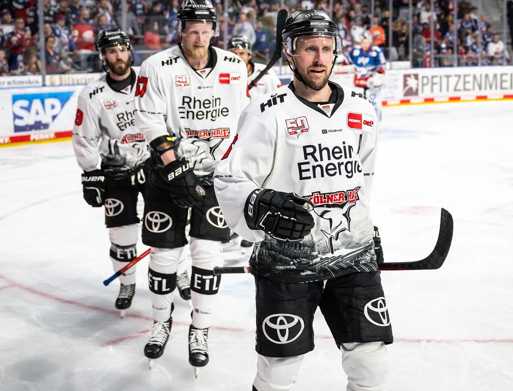 Tres jugadores de hockey sobre hielo con camisetas blancas con logotipos negros y diseños gráficos están de pie sobre el hielo.