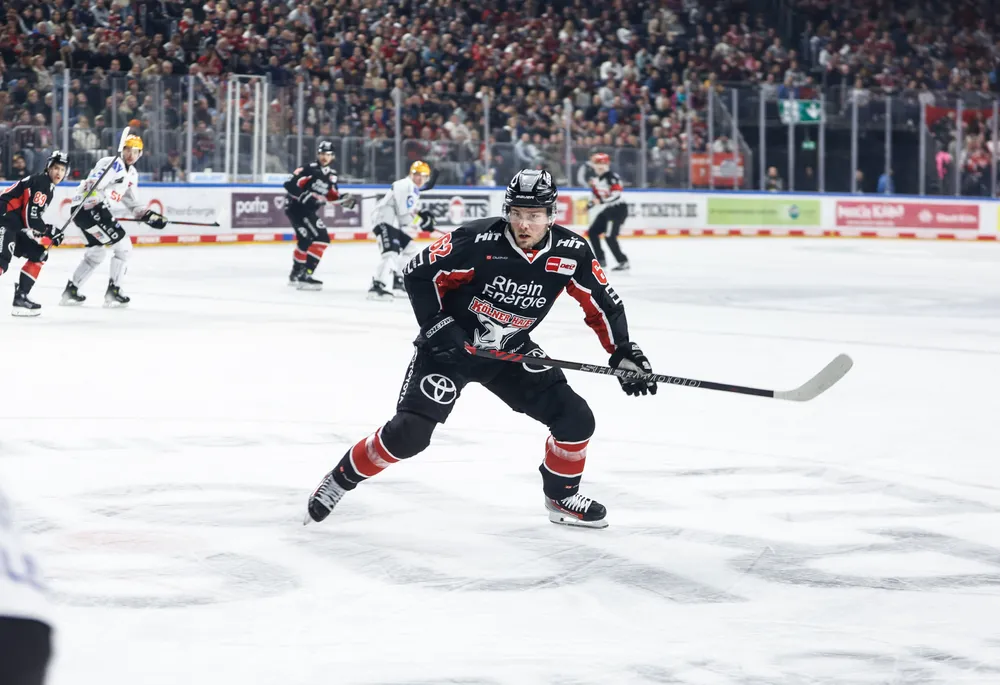 Un jugador de hockey sobre hielo con una camiseta negra con acentos rojos y blancos, patinando sobre el hielo con un palo.