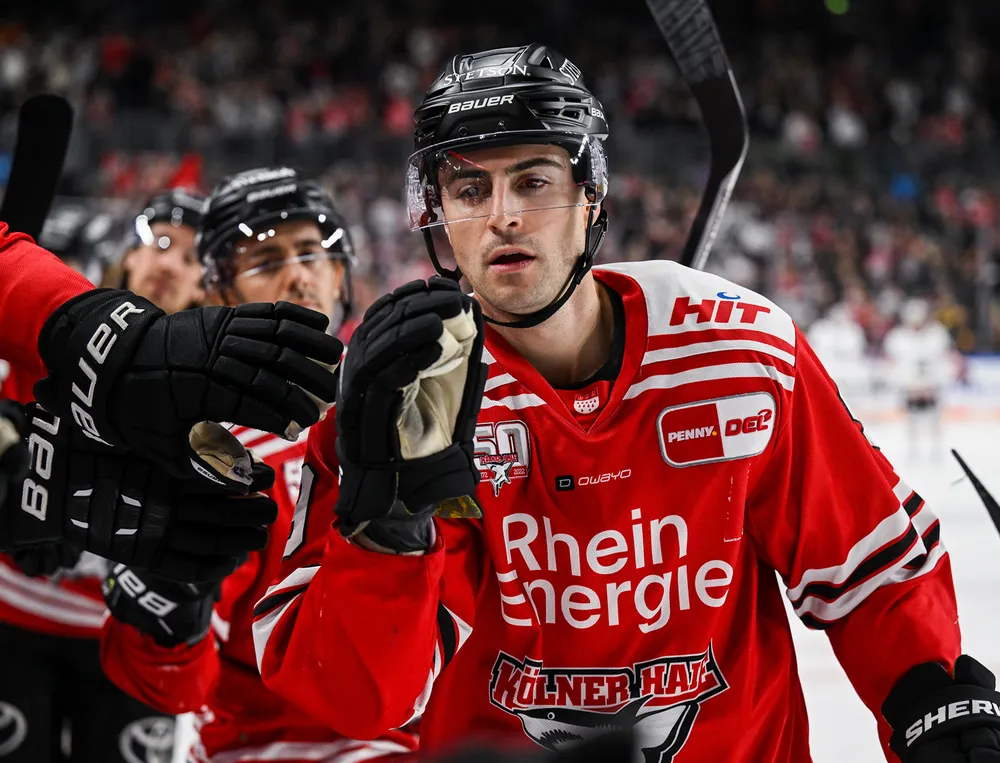 Un jugador de hockey lleva una camiseta roja con detalles en blanco y negro mientras interactúa con un compañero de equipo.