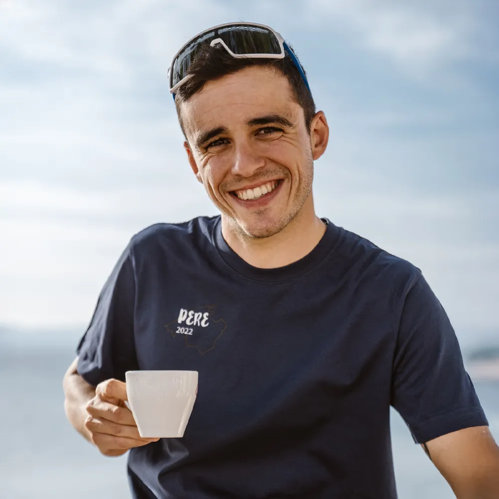 Un hombre sostiene una taza y lleva una camiseta azul marino con la impresión 'PERE 2022', sonriendo frente a un fondo borroso.