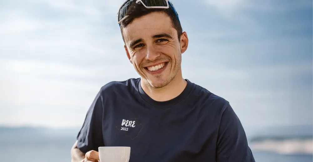 Un hombre atlético con una camiseta azul marino que dice 'PERE 2022' sonríe mientras sostiene una taza, con un fondo costero borroso.