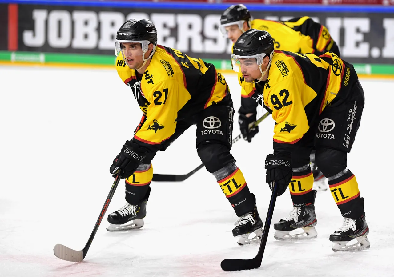 Dos jugadores de hockey sobre hielo con uniformes amarillos y negros con los números 21 y 92 en la pista de hielo, concentrados en el juego.