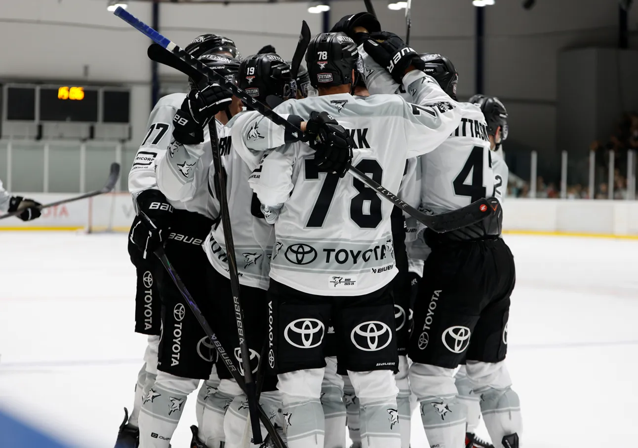 Jugadores de hockey sobre hielo con uniformes blancos con números negros y logos de patrocinadores, abrazándose en la pista de hielo.