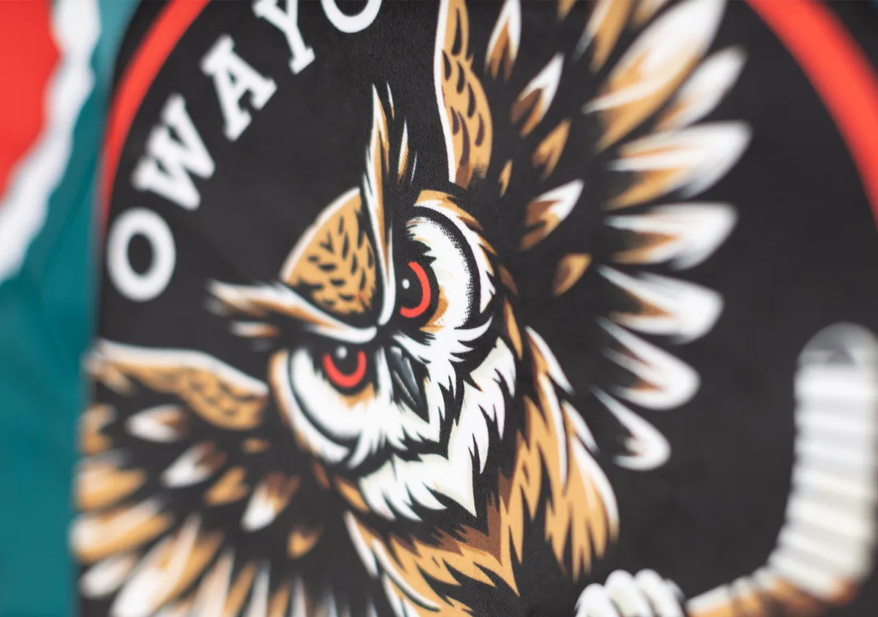 Una cabeza de búho estilizada con ojos rojos sobre fondo negro, rodeada por un círculo rojo y blanco, en una prenda deportiva de owayo.