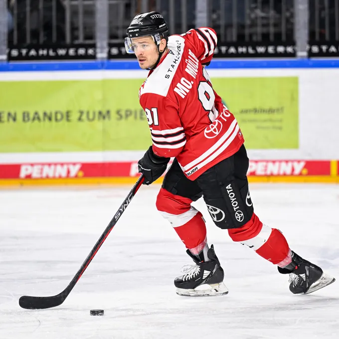 Un jugador de hockey sobre hielo con un jersey rojo y negro con el número 91 y el nombre Mc. Miller en la espalda, de pie sobre el hielo y controlando un disco con su palo.