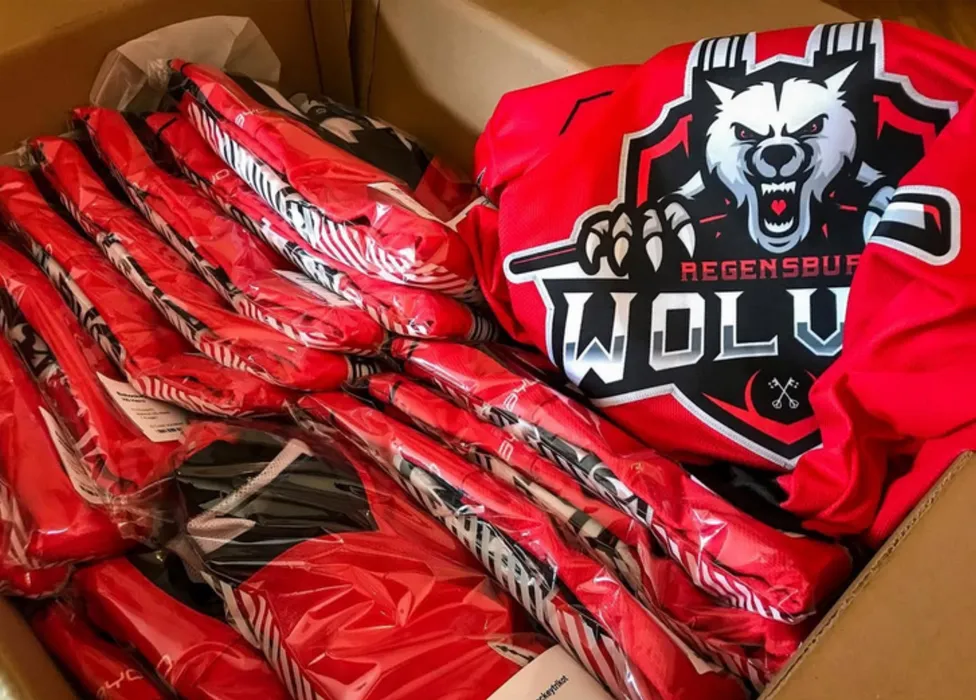 Una caja llena de camisetas deportivas rojas personalizadas, incluyendo una con un logo de lobo y la inscripción 'Regensburg Wolves'.