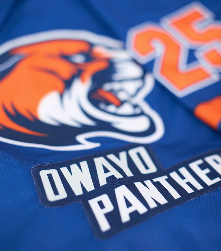 Acercamiento de una camiseta deportiva azul con un logotipo de pantera naranja y la inscripción 'OWAYO PANTHERS' en letras blancas sobre una etiqueta oscura.