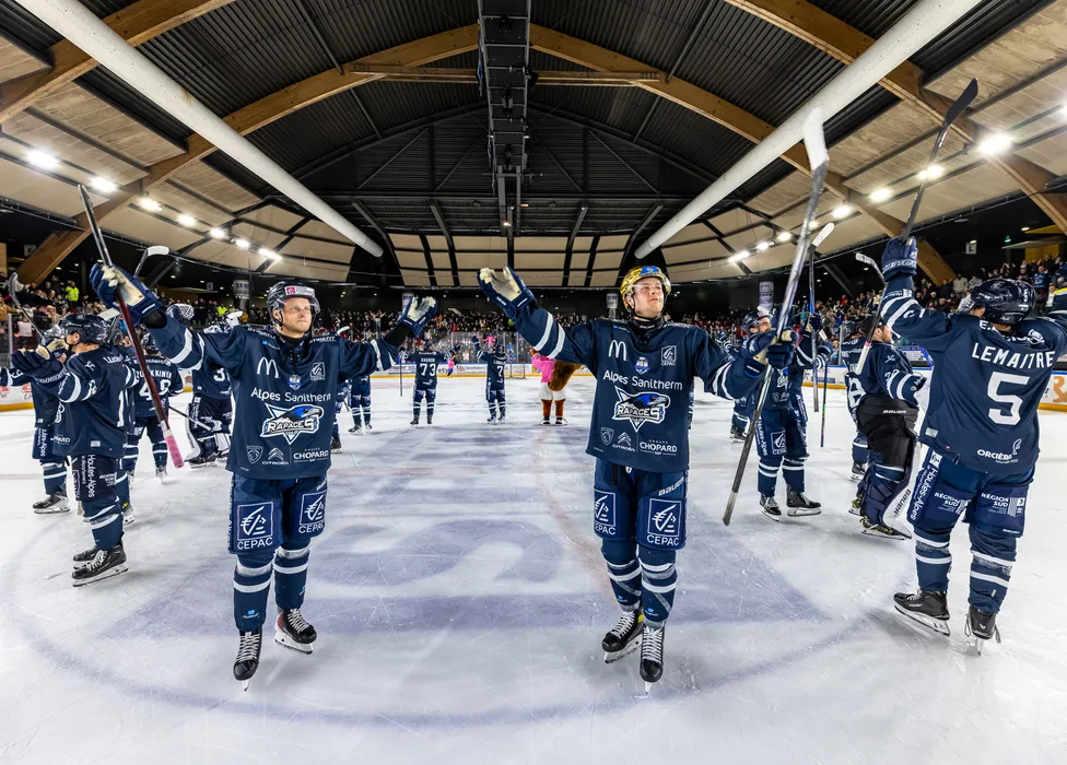 Jugadores de hockey sobre hielo con camisetas azules están en el hielo, celebrando con los palos en alto frente a una multitud animada.