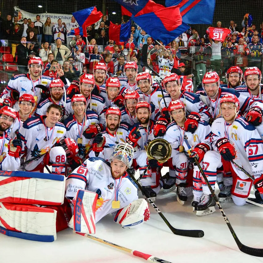 Un equipo de hockey posa orgullosamente sobre el hielo con medallas y un trofeo mientras los aficionados vitorean al fondo.