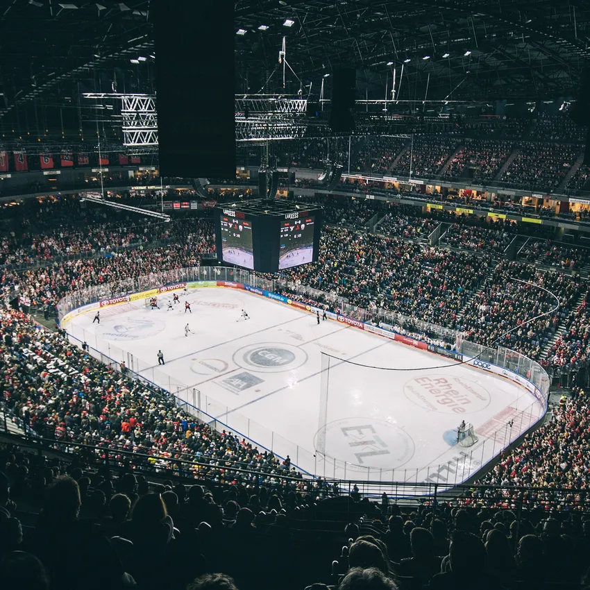 Una arena de hockey sobre hielo llena durante un partido, con jugadores en el hielo y un gran marcador de fondo.