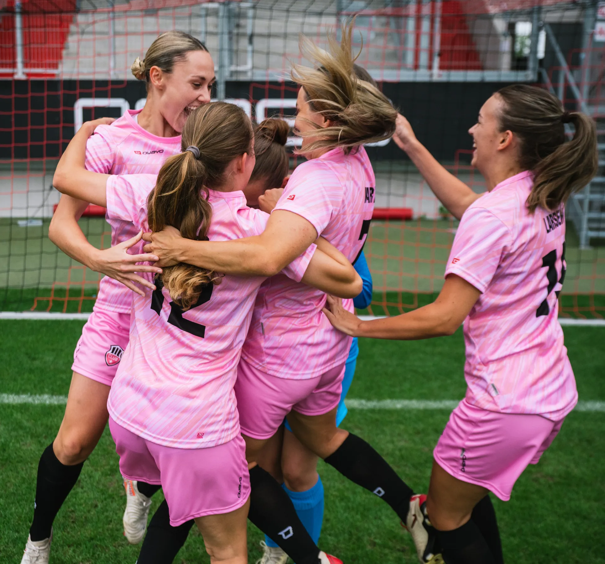 Cinco jugadoras en camisetas rosas celebran juntas con alegría en el campo de fútbol.