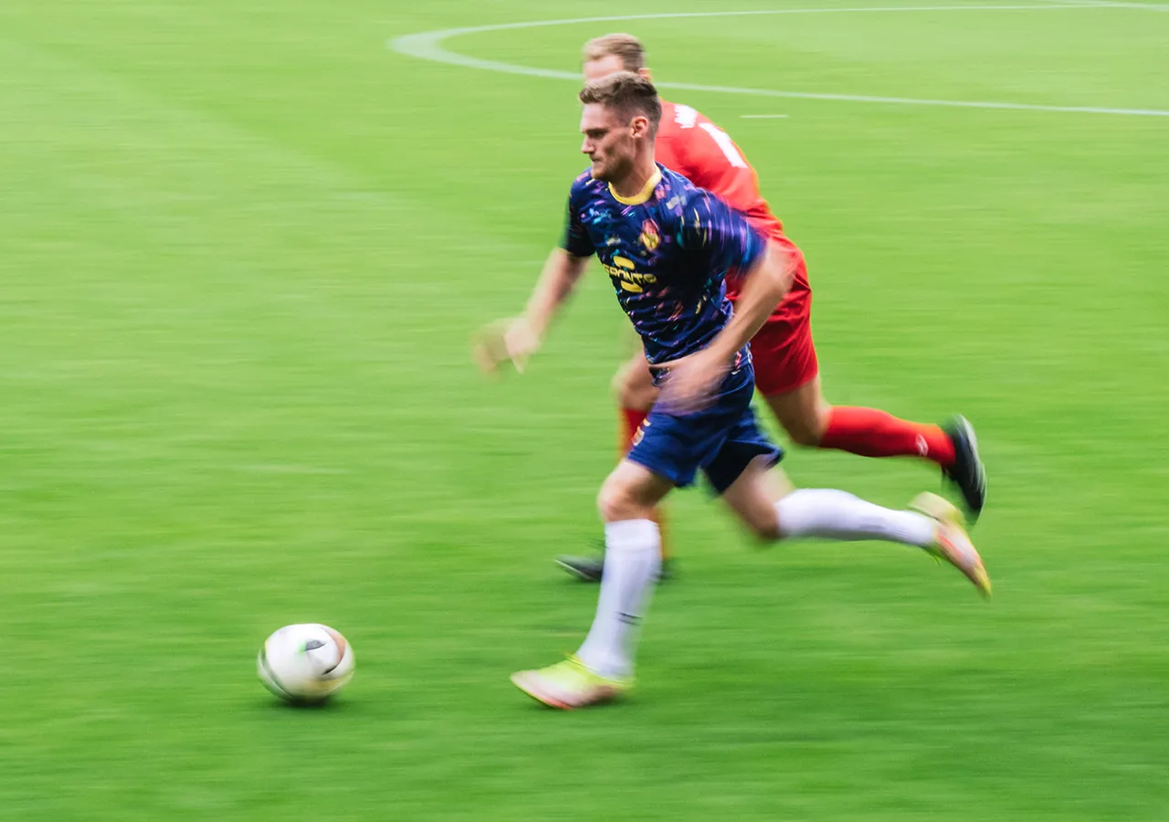 Un jugador de fútbol con una camiseta colorida corre por el césped mientras otro jugador lo sigue de cerca.