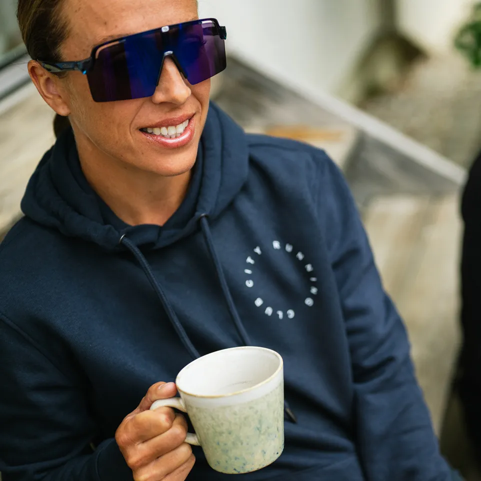 Una persona con una sudadera azul oscura con un logo sonríe a la cámara mientras sostiene una taza.