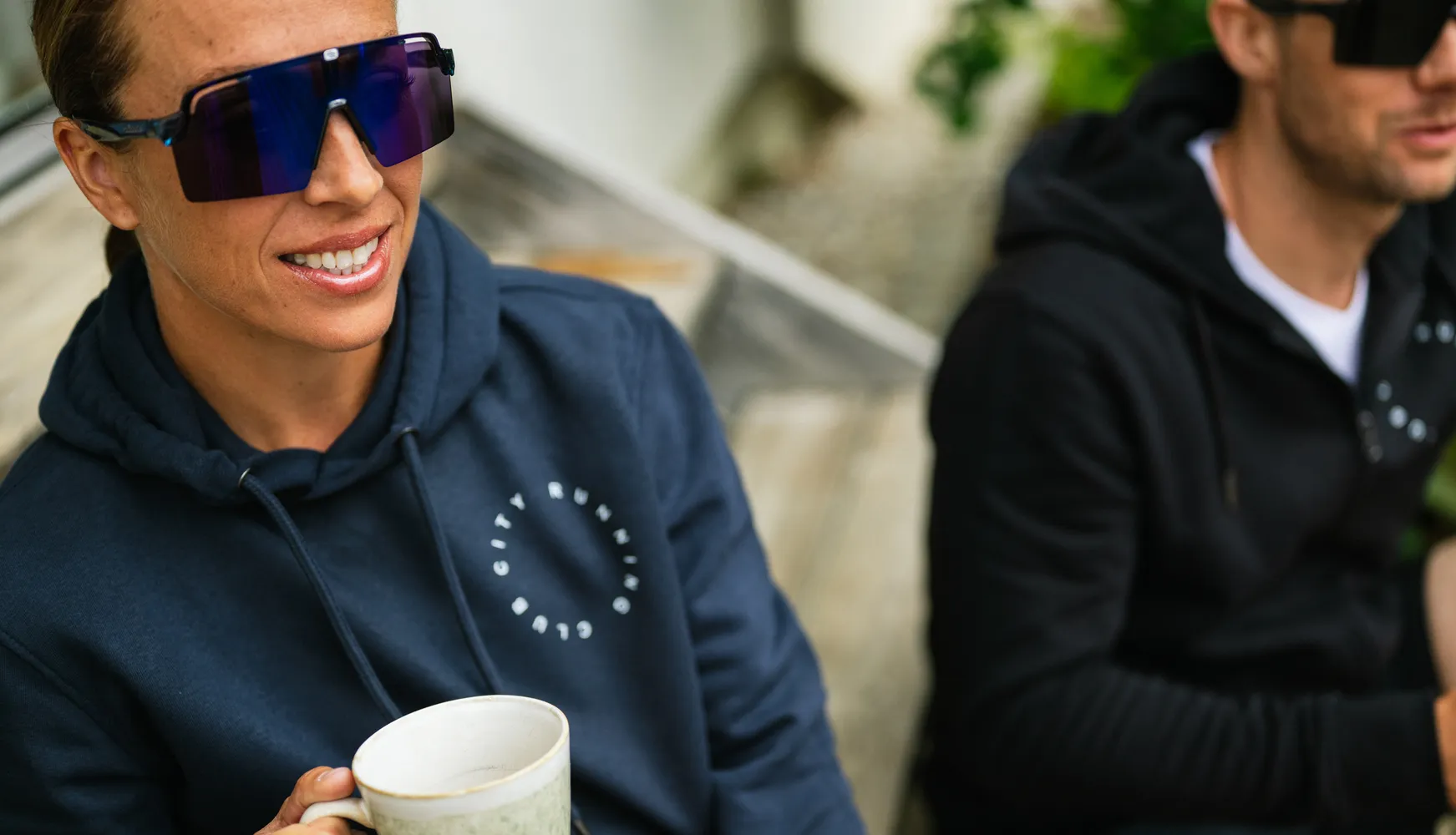 Una persona con una sudadera azul sostiene una taza y lleva grandes gafas de sol, mientras que otra persona en el fondo también lleva una sudadera.