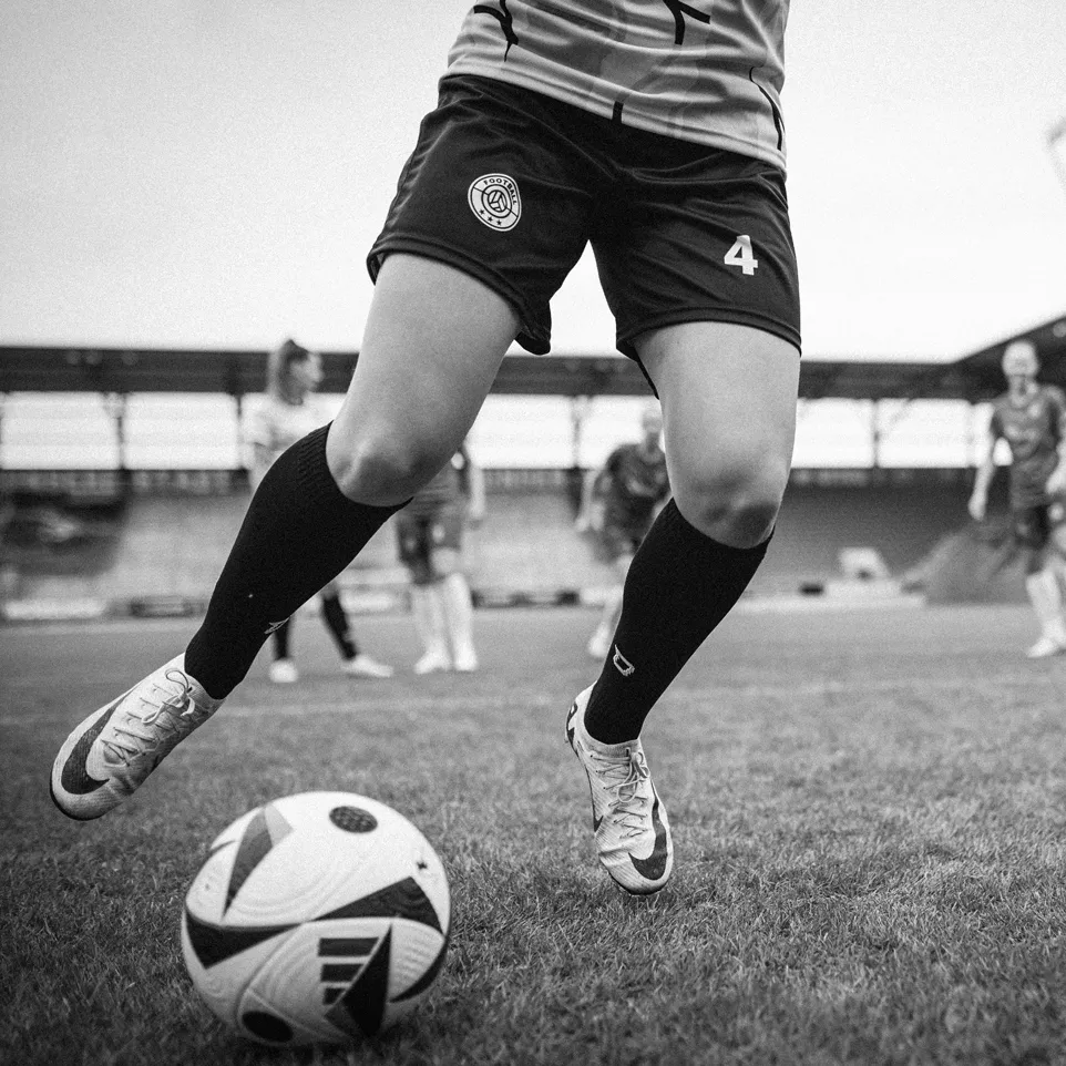Una jugadora con pantalones cortos y medias driblando un balón de fútbol en un campo de hierba.