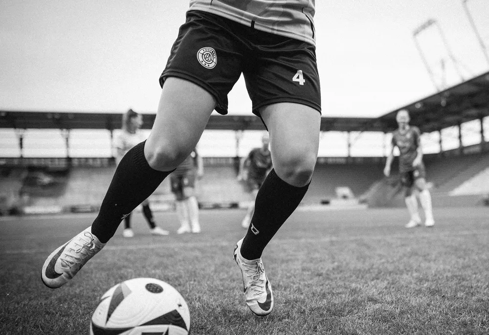 Una jugadora con pantalones cortos de fútbol y calcetas largas está driblando un balón en el campo.