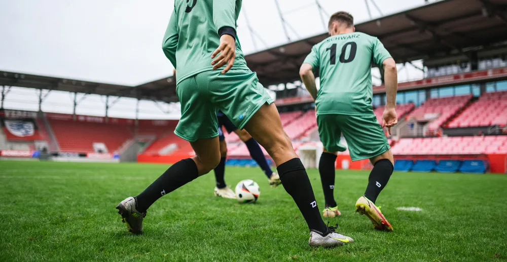 Dos futbolistas con camisetas verde claro y calcetines negros entrenando en un campo de hierba en un estadio.