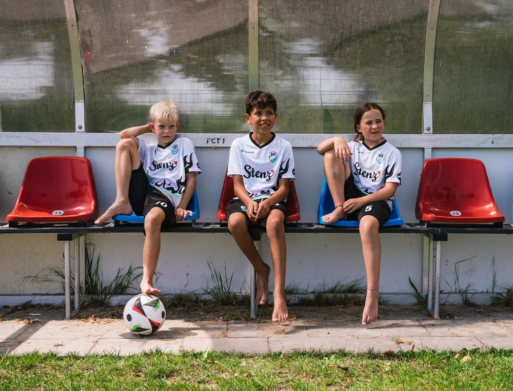Tres niños están sentados en un banco al borde de un campo de fútbol, vistiendo camisetas de fútbol personalizadas con un balón de fútbol frente a ellos.