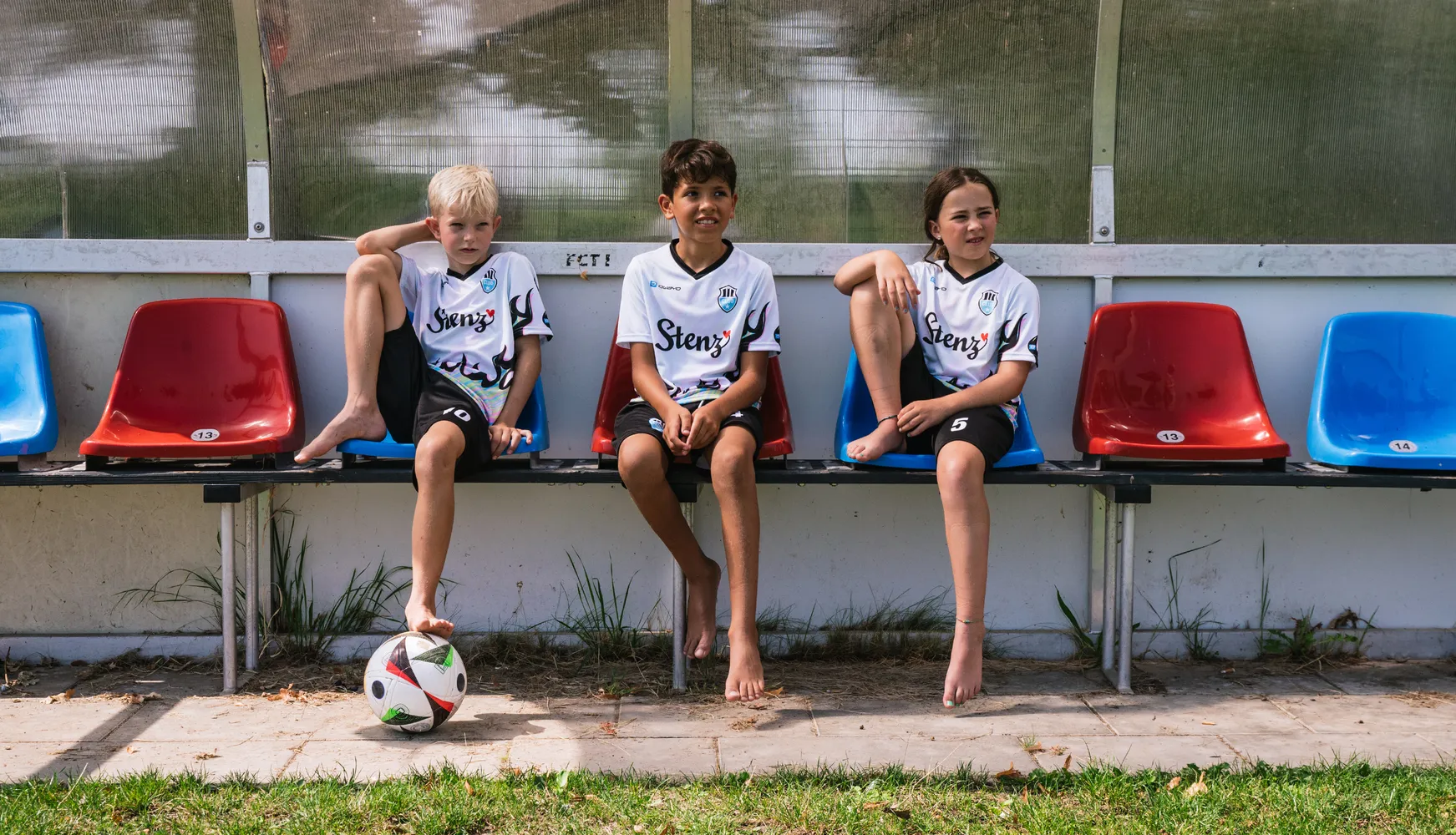 Tres niños con camisetas de fútbol personalizadas están sentados en un banco con un balón de fútbol en el suelo.