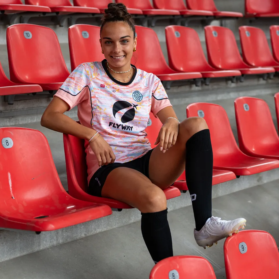 Una mujer sonriente está sentada en un estadio sobre un asiento rojo, vestida con una camiseta de fútbol colorida y calcetines negros.