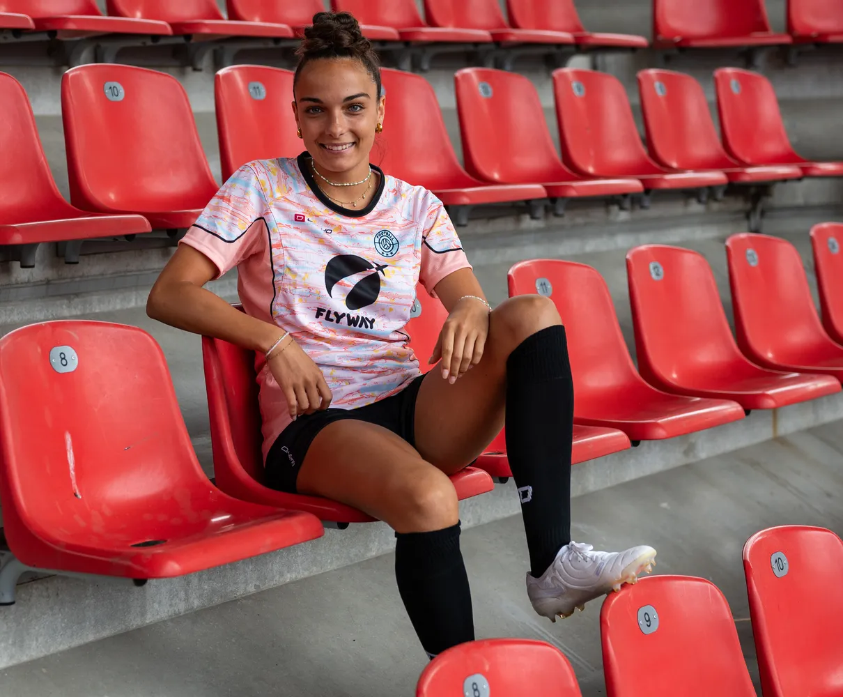 Una joven está sentada en un asiento de estadio rojo, vistiendo una camiseta de fútbol colorida y calcetines negros.