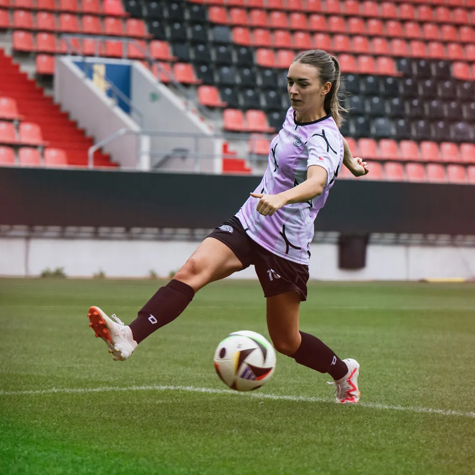 Una jugadora patea un balón de fútbol en un campo de estadio, vestida con una camiseta morada y pantalones cortos negros.