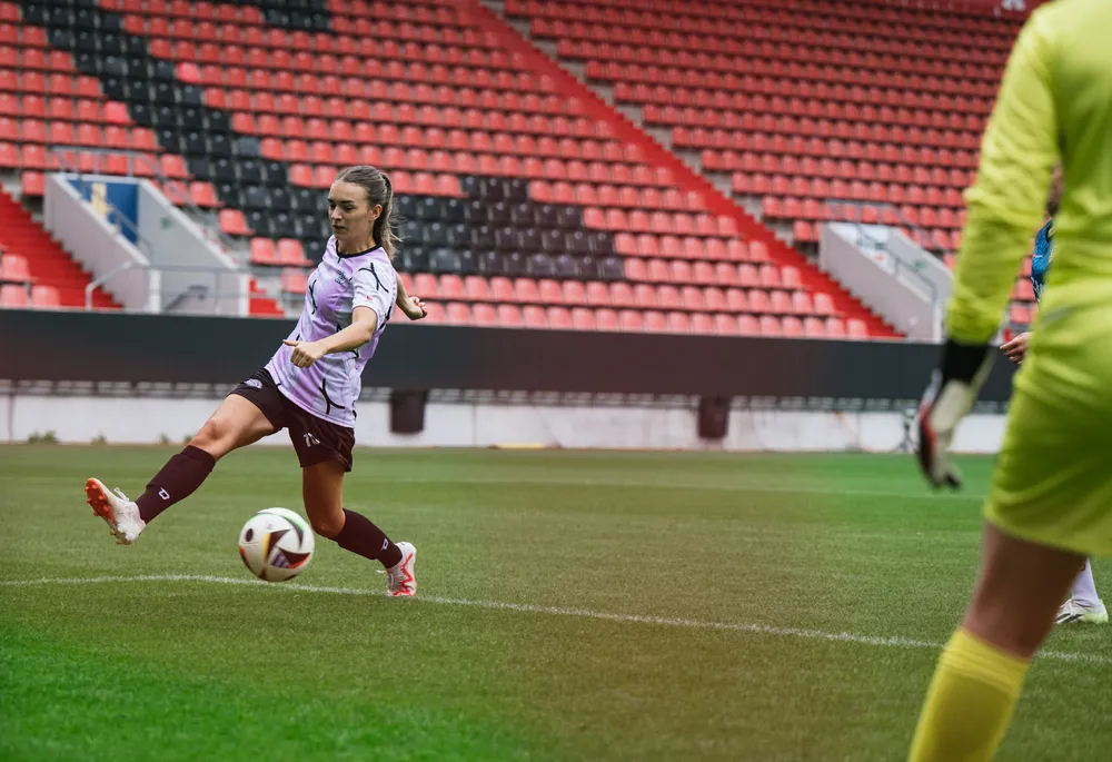 Una futbolista con una camiseta morada y negra patea un balón en un campo con gradas vacías al fondo.