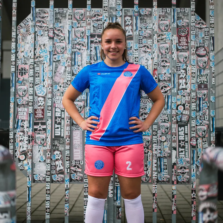 Una niña sonríe frente a una pared cubierta de pegatinas, vistiendo una camiseta de fútbol azul con una franja rosa y pantalones cortos rosas.