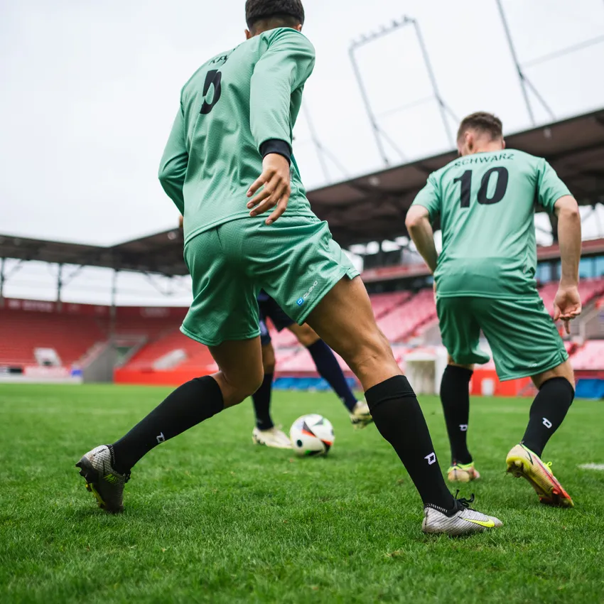 Dos futbolistas con camisetas verdes y calcetas negras jugando en un campo de césped con un balón.