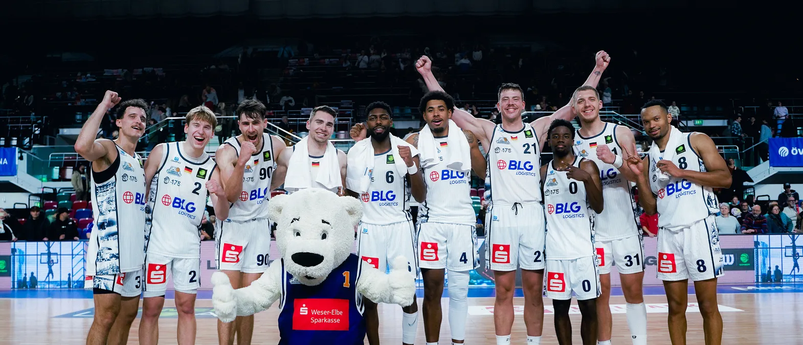 Jugadores de baloncesto en camisetas blancas con el logo de BLG Logistics celebran en la cancha, rode por una multitud, con una mascota oso polar en primer plano.