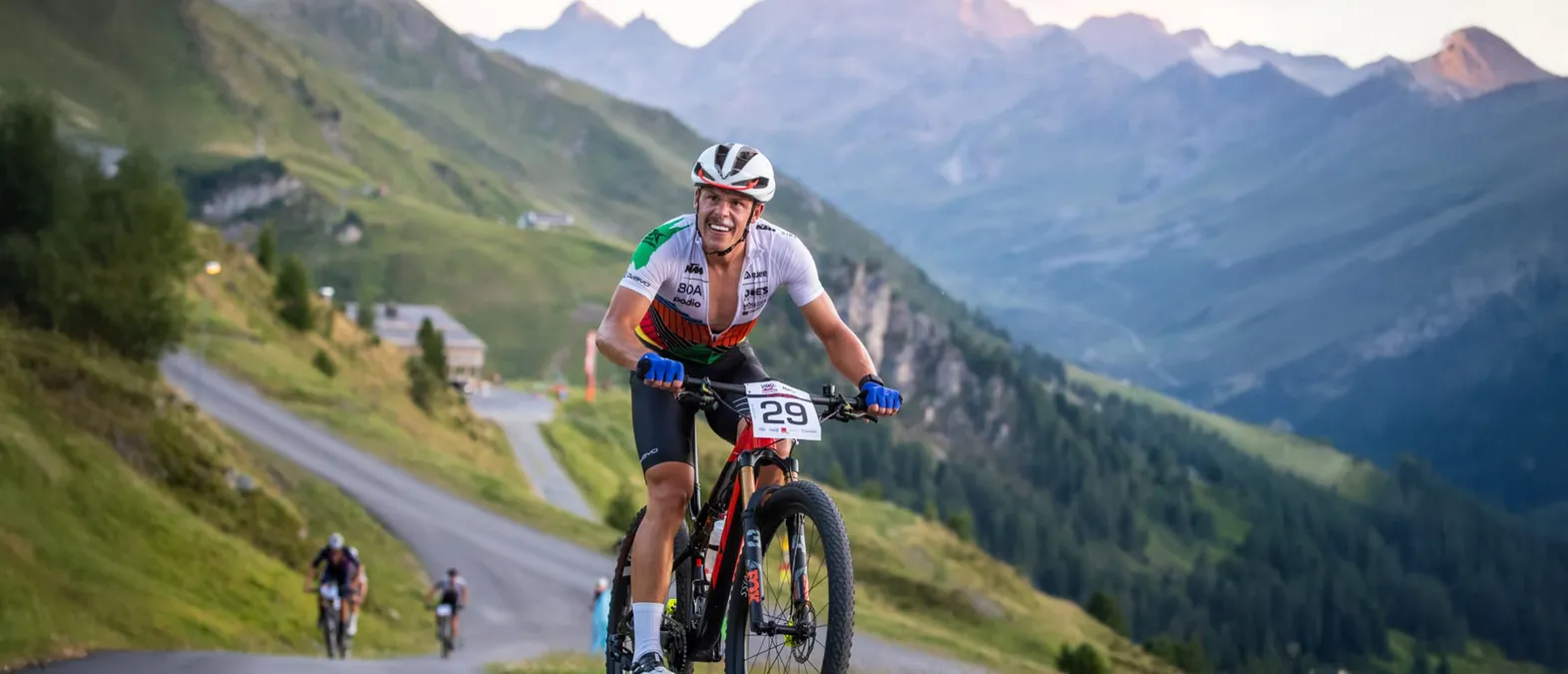 Un ciclista con un maillot blanco, verde y naranja con el número 29 pedalea por una carretera de montaña en un paisaje montañoso.