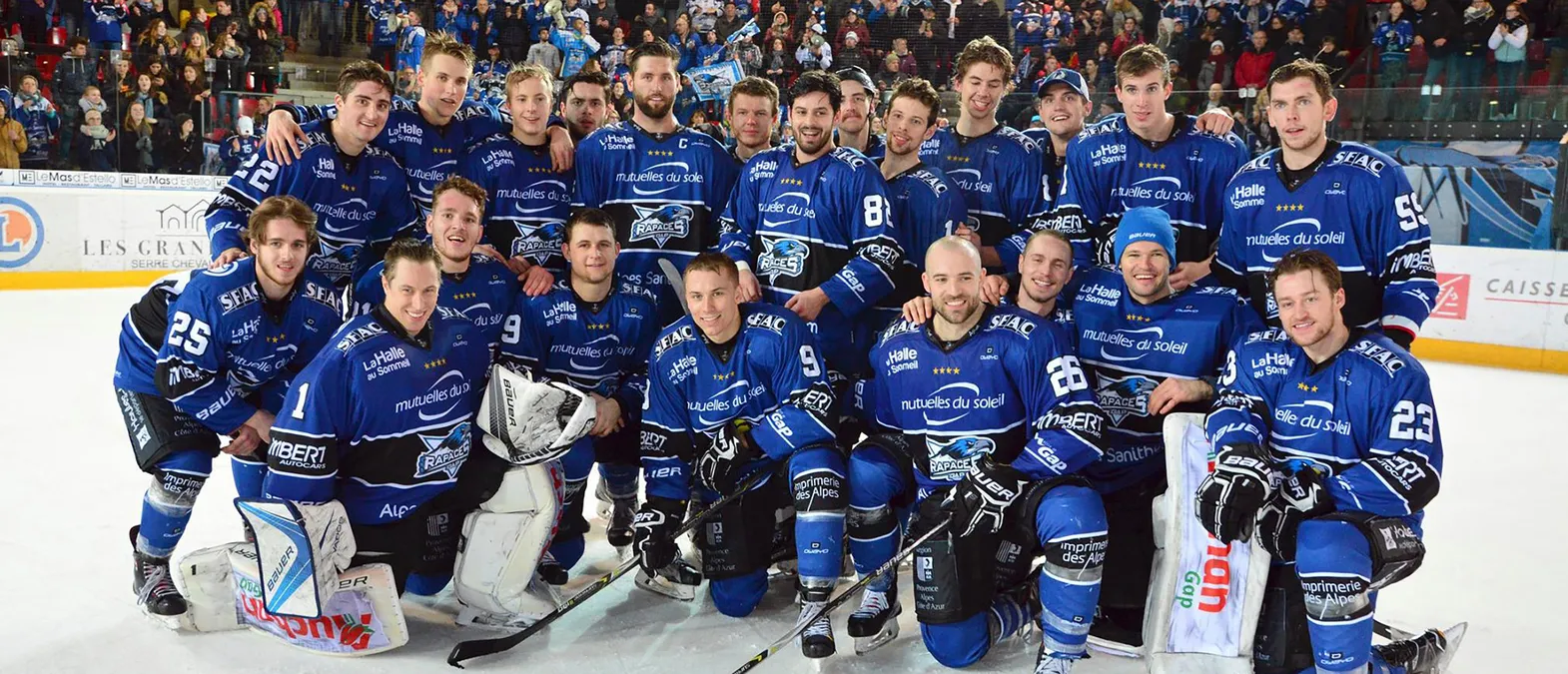 Un equipo de hockey sobre hielo con uniformes azules y el logo de los Rapaces de Gap posa en la pista frente a una multitud.