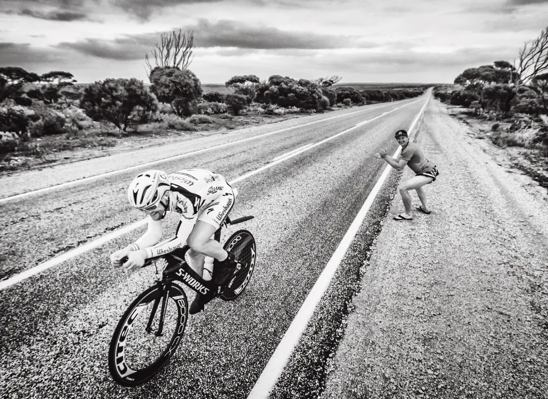 Un ciclista en posición aerodinámica en una carretera, mientras una persona al lado de la carretera hace señas.