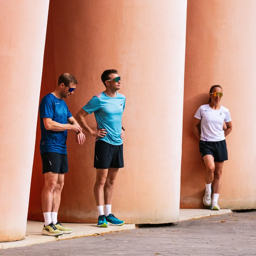 Tres atletas en ropa deportiva y gafas de sol están apoyados de manera casual contra columnas rosas.