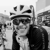 Un ciclista sonriente con una chaqueta de ciclismo negra con la inscripción 'BIKE INDUSTRIES' en la manga y un casco blanco, usando gafas de sol que reflejan el entorno.