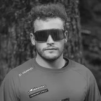 Un hombre con cabello rizado y gafas de sol lleva una camiseta deportiva gris de manga larga con el logo 'owayo' en el hombro y otros logos de patrocinadores en el pecho.