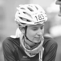 Una ciclista con un maillot de ciclismo negro con el logo 'IWC Schaffhausen', un casco blanco con el número 183 y una bufanda alrededor del cuello.