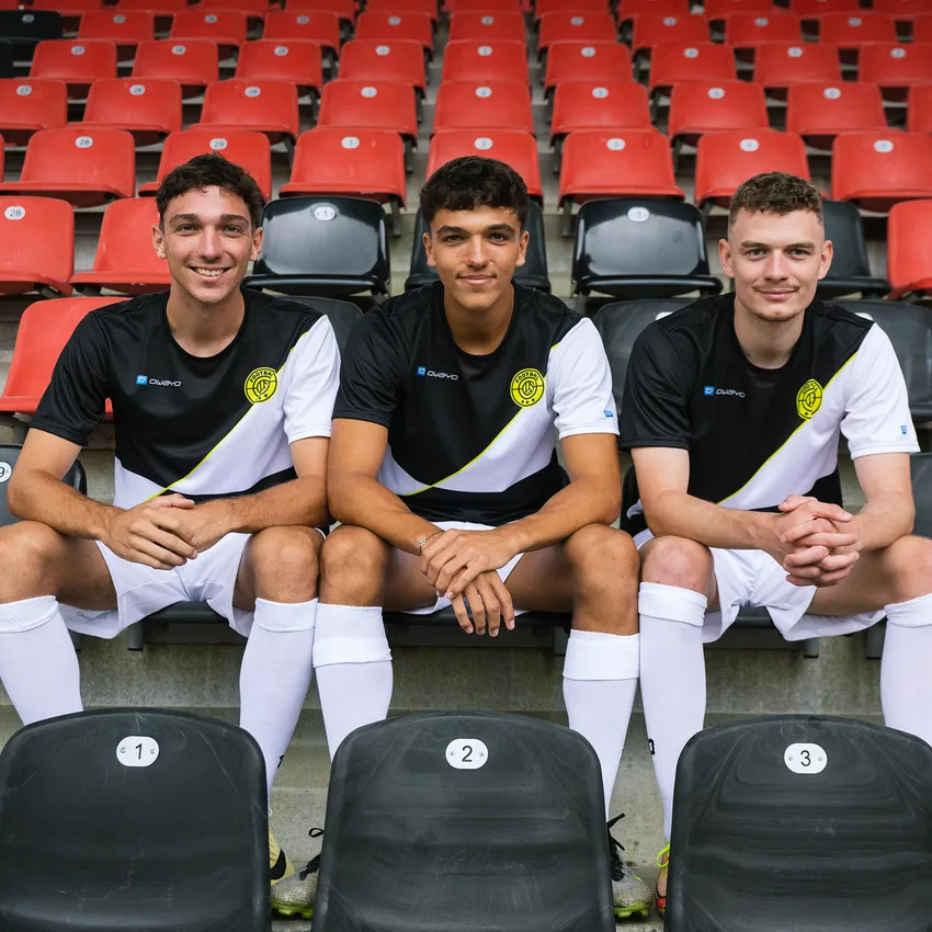 Tres jóvenes en ropa deportiva de owayo se sientan en un estadio sobre asientos numerados frente a gradas rojas.