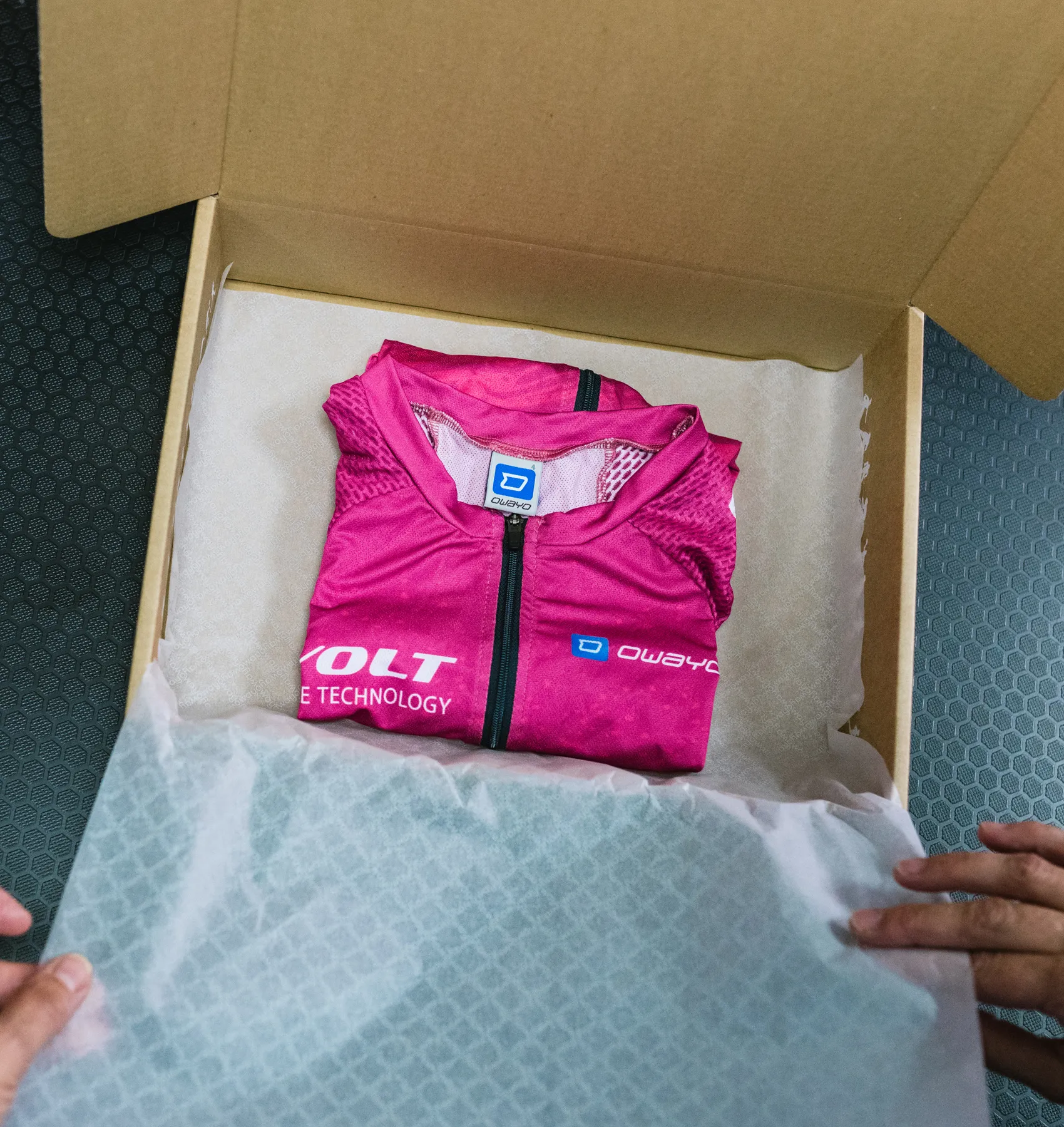 Una prenda deportiva personalizada de color rosa de owayo, doblada, está dentro de una caja de cartón abierta sobre papel blanco, sostenida por manos.