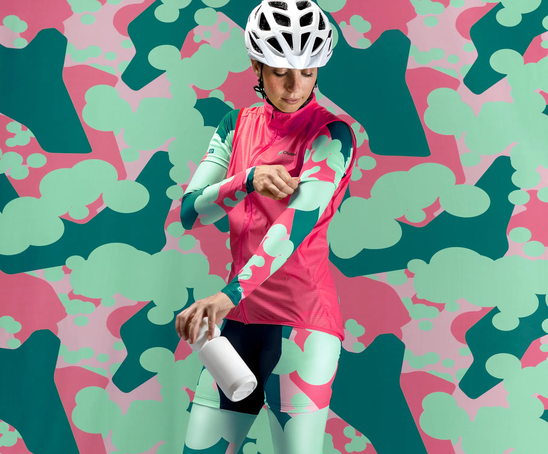 Una ciclista con ropa deportiva personalizada de color rosa y verde con un patrón abstracto, ajustando sus mangas mientras sostiene una botella blanca, frente a un fondo colorido con el mismo patrón.