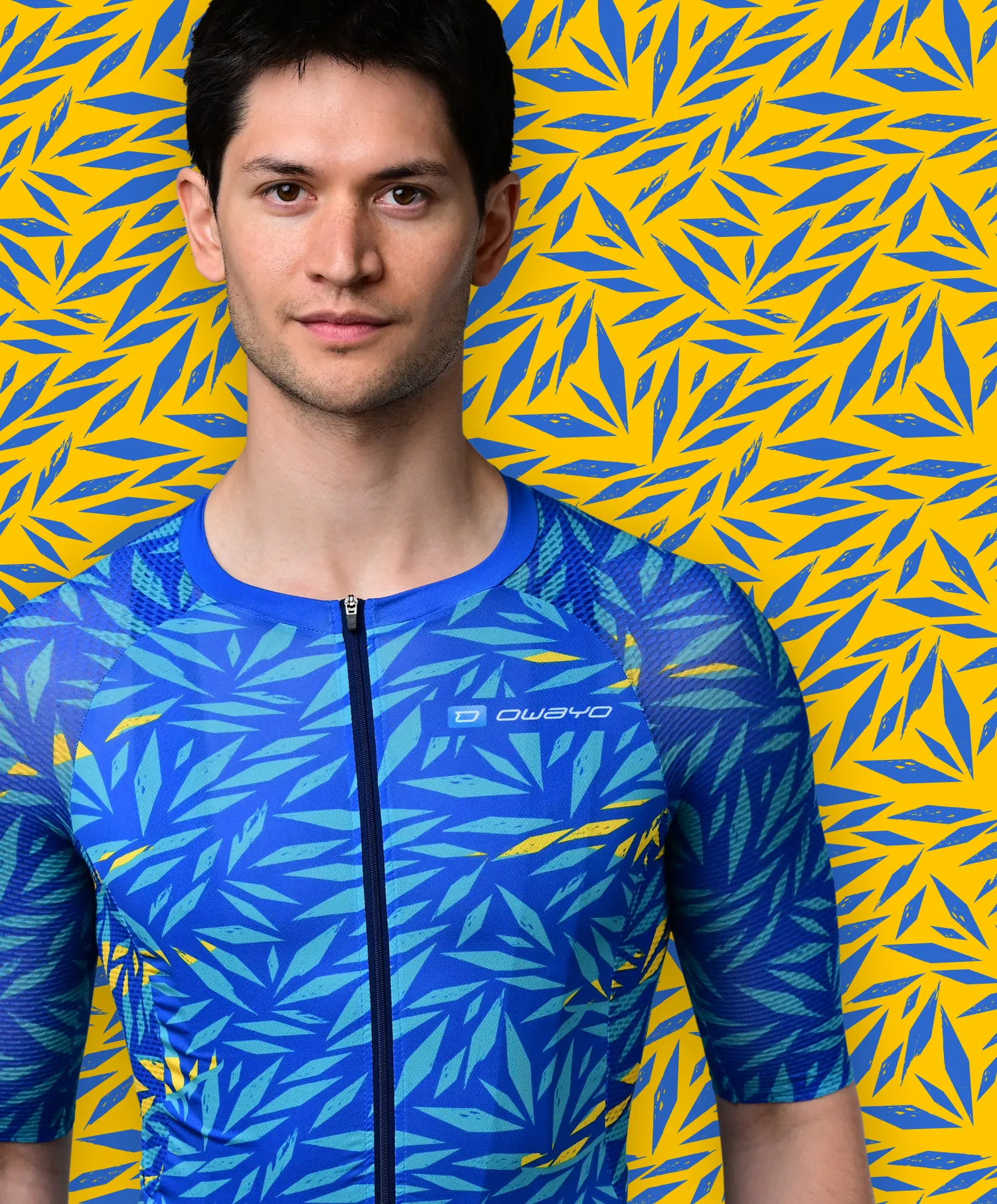 Un hombre lleva una camiseta deportiva azul de manga corta con un patrón de hojas azules y amarillas que recuerda a una planta tropical, frente a un fondo amarillo con patrones de hojas azules.