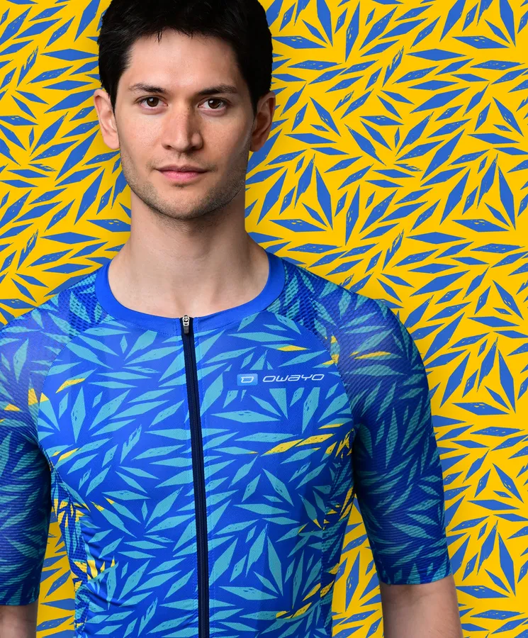 Un hombre lleva una camiseta deportiva azul de manga corta con un patrón de hojas azules y amarillas que recuerda a una planta tropical, frente a un fondo amarillo con patrones de hojas azules.