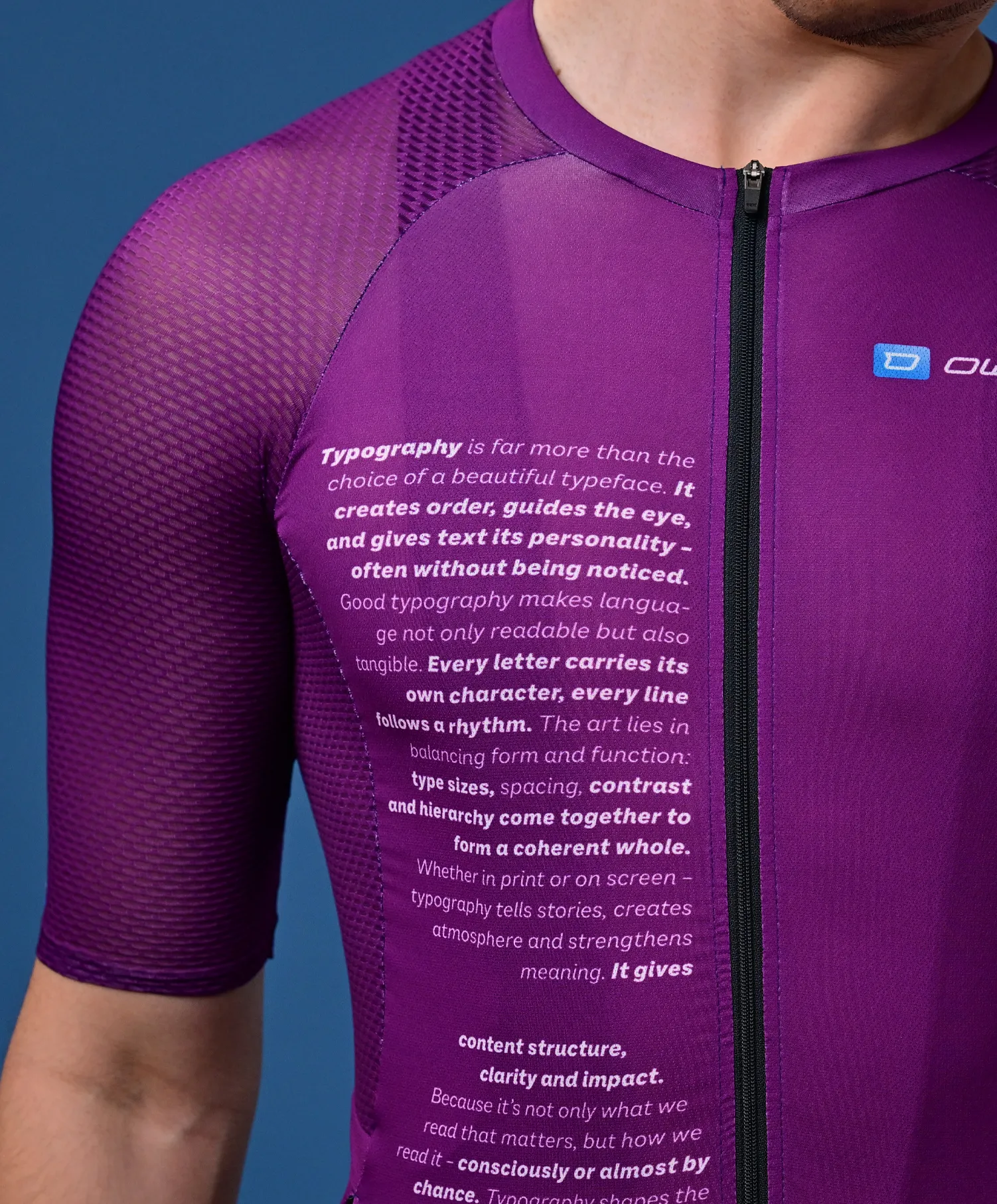 Un hombre lleva una camiseta deportiva morada de manga corta con un texto sobre tipografía en el pecho y un pequeño logo en el hombro.